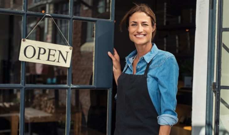 Portrait of a happy waitress standing at restaurant entrance. Portrait of mature business womanattend new customers in her coffee shop. Happy woman owner showing open sign in her small business shop.
